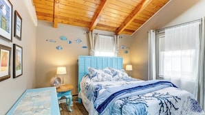 Een slaapkamer met een houten plafond, een bed met blauw beddengoed, een nachtkastje met een lamp en een klein tafeltje met een fotolijst.
