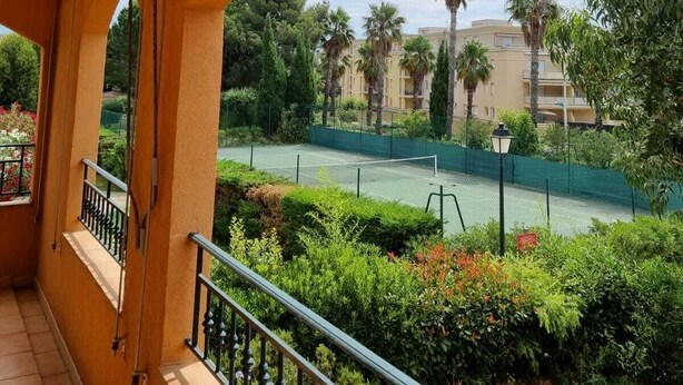 Un balcó amb vista a una pista de tennis i vegetació circumdant, una barana i un edifici al fons.