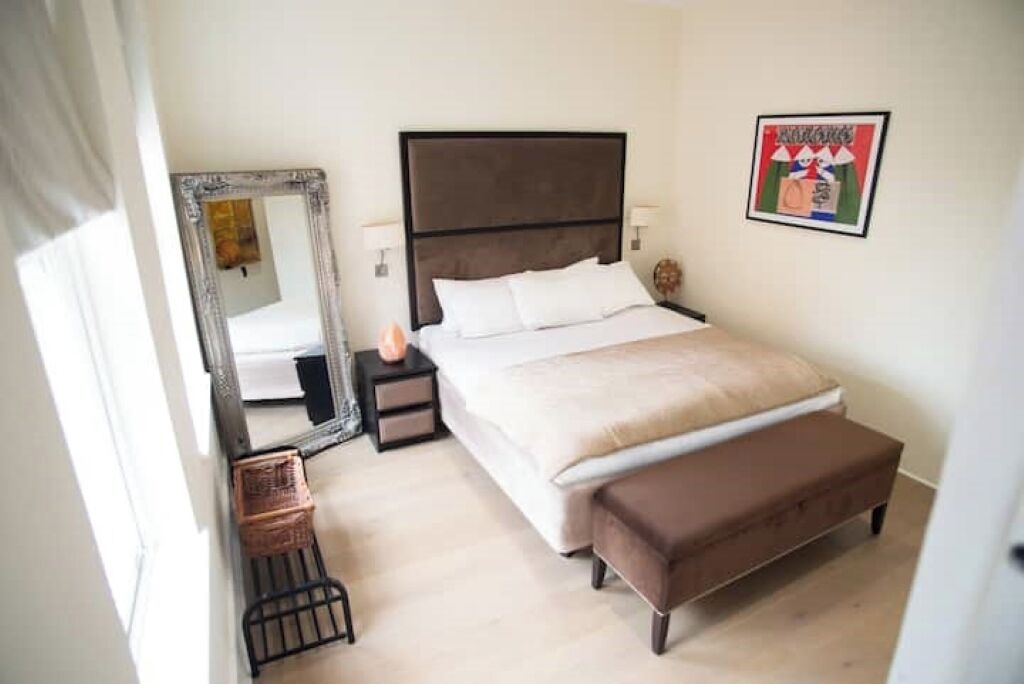 Quarto com uma cama grande, uma mesa de cabeceira, um espelho, uma obra de arte na parede e um pequeno banco.