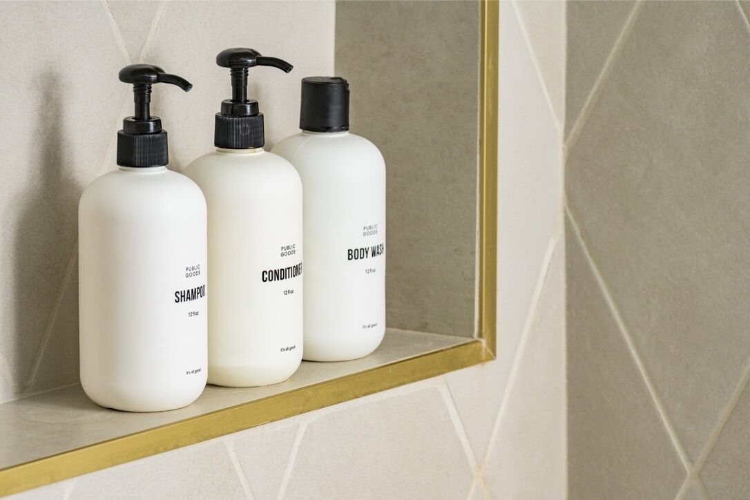 Drei weiße Flaschen von Public Goods Shampoo, Conditioner und Duschgel auf einem Regal.