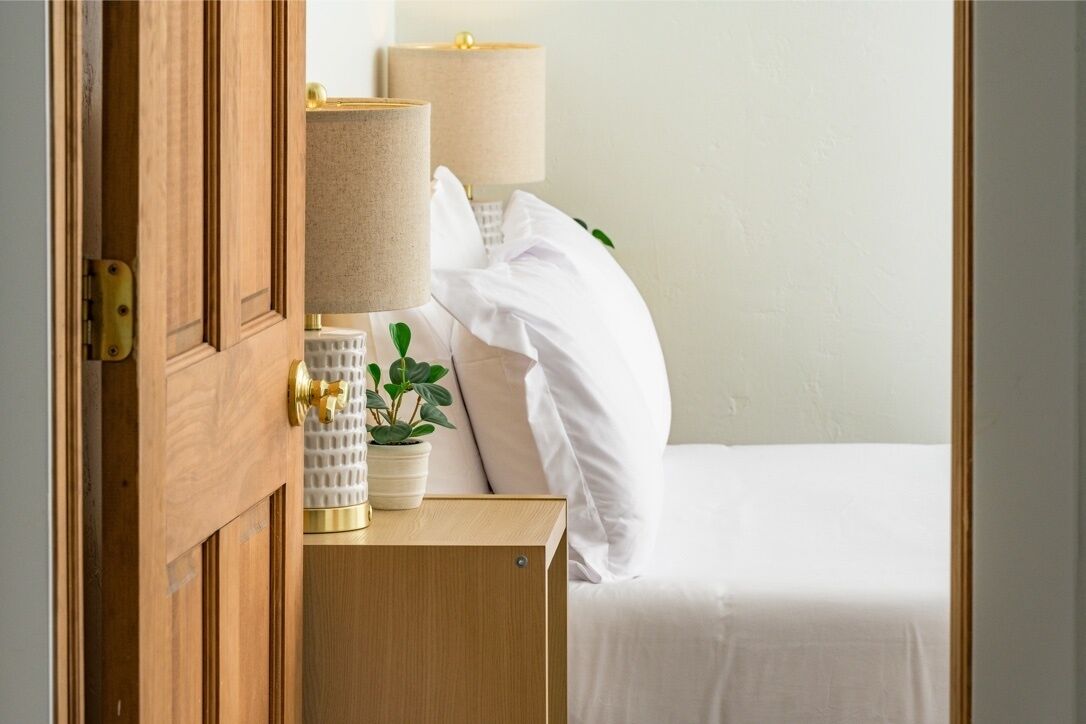 Una cama bien hecha con ropa blanca, una lámpara de noche, una pequeña planta en maceta y una mesita de noche de madera.