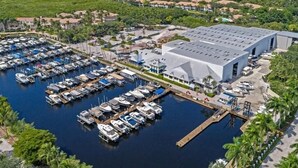 Une vue aérienne d’une marina où sont amarrés de nombreux bateaux, d’un grand bâtiment doté d’un toit plat et des quartiers résidentiels environnants, parsemés de palmiers.