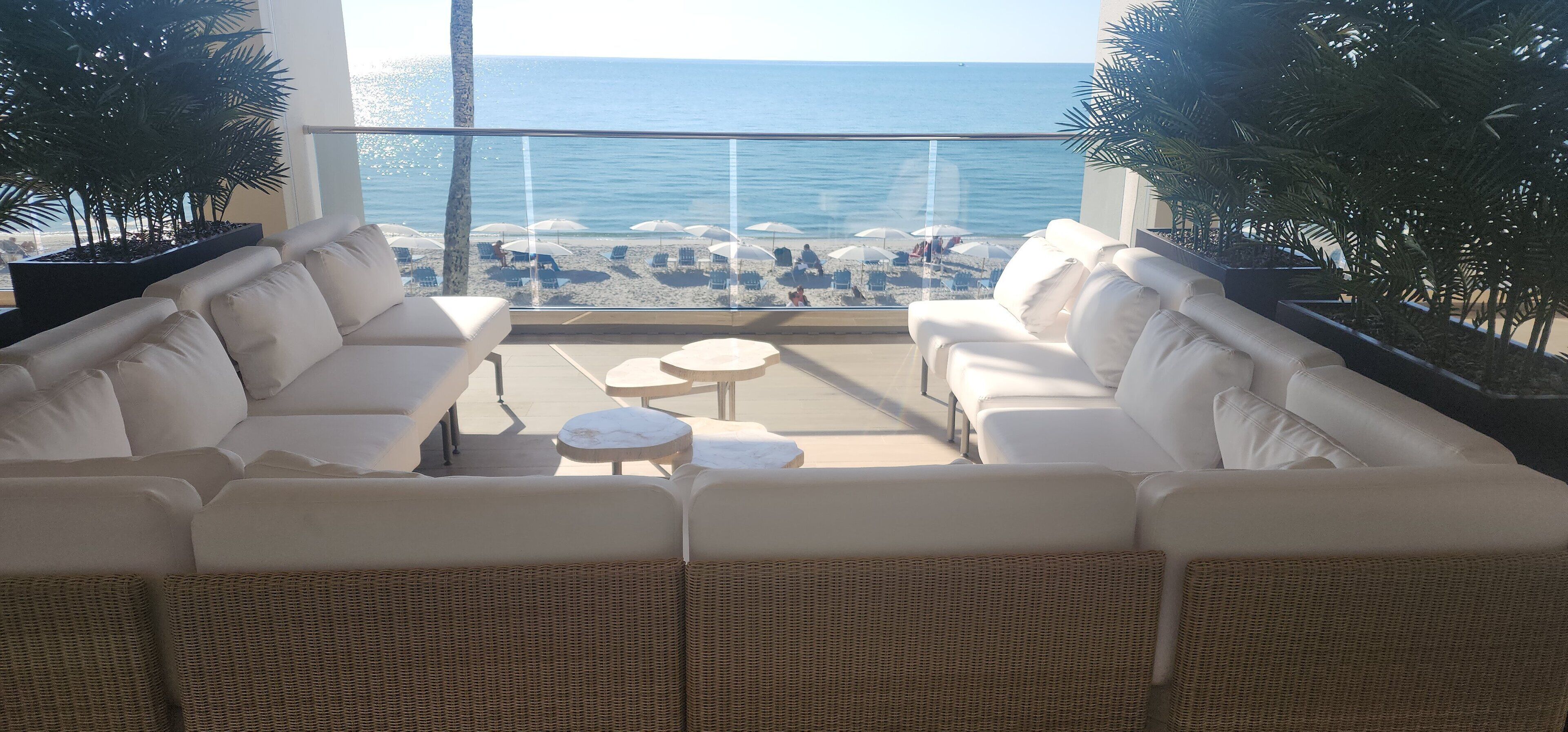 A balcony with a white sofa and two round tables, overlooking a beach with umbrellas and people.