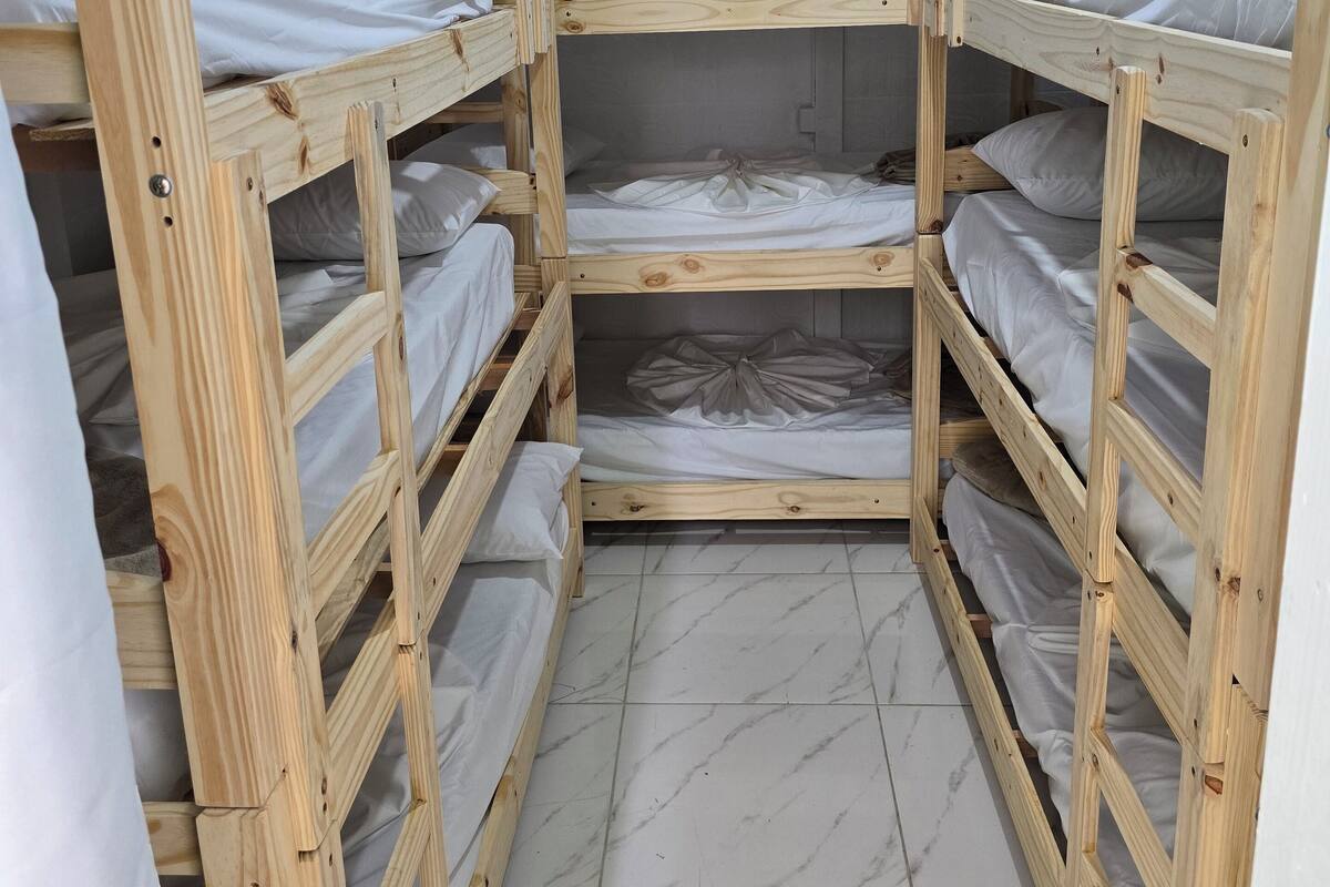 A corner room with four wooden bunk beds, each with white bedding. The room has a tiled floor and a plain wall.