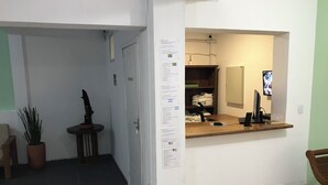 Un área de recepción con mostrador, una planta en maceta, una pequeña mesa con un adorno y una pantalla montada en la pared.