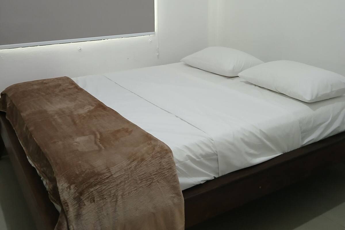 A single bed with white bedding and a brown blanket, a window with a blind, and a plain white wall.