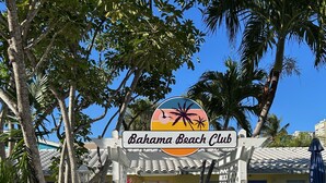 Entrada del Bahama Beach Club amb un camí que condueix a l'edifici, envoltat de palmeres i una tanca blanca.