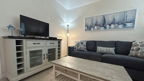 Ein Wohnzimmer mit Fernseher auf einer Kommode, einer Couch mit gemusterten Kissen und einem Segelbootbild an der Wand.