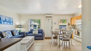 Ein Wohnzimmer mit Sofa, Couchtisch und Esstisch mit Stühlen. Eine Küchenzeile mit Schränken und Geräten ist ebenfalls vorhanden.