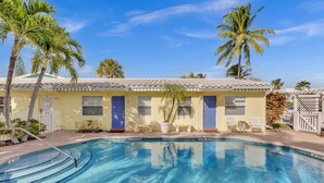 A poolside view of a yellow building with a blue door, surrounded by palm trees and a clear blue sky.