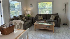 A living room with two sofas, a coffee table, a white chair, and a floor lamp. There are windows with blinds, a ceiling fan, and a wall-mounted light fixture.