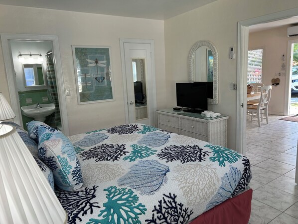 A bedroom with a bed, a television on a dresser, a bathroom with a sink and mirror, and a dining area with chairs and a table.