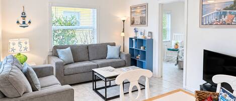 A modern living room with a grey sofa, a coffee table, and a TV. There is a wicker basket with snacks and a sign that says 'Relax Explore Make Memories Welcome!'.