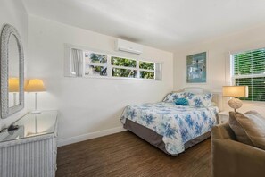 Une chambre à coucher comprenant un lit, une table de chevet, une lampe, un miroir, une fenêtre avec des rideaux et un climatiseur fixé au mur.