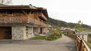 Un bâtiment traditionnel en bois, doté d’une façade en pierre, d’un balcon orné de sculptures complexes et d’une allée pavée menant à un village.