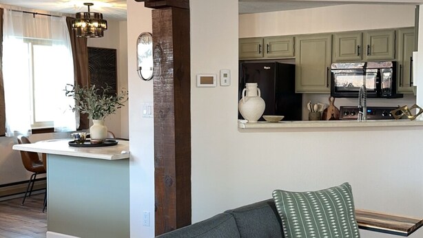 Un soggiorno con un divano grigio, due cuscini decorati, una ventola a soffitto in legno e una zona cucina sullo sfondo.