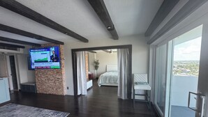 A modern hotel room with a large TV on the wall, a balcony with a view, and a separate bedroom area.