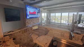 A modern kitchen with a marble countertop, a large window with a city view, and a mounted television displaying financial news.