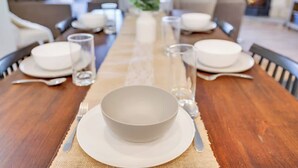 Una mesa de comedor dispuesta con platos, tazones, vasos y cubiertos, ubicada en el centro de una habitación con sofás y una chimenea al fondo.