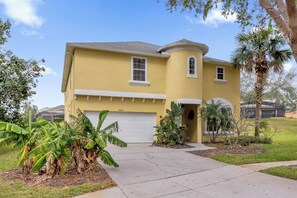 A yellow house with a garage, surrounded by tropical plants and a clear sky. - Pool, Themed Rooms, Resort (Kissimmee)