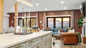 A modern kitchen with a large island, a dining area with a sofa and table, and a view of mountains through large windows.