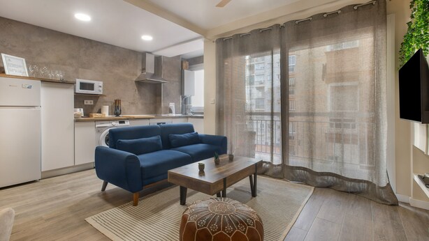Una sala de estar moderna con sofá, mesa de centro y reposapiés, un área de cocina con electrodomésticos y un amplio ventanal con cortinas.