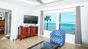 Una sala de estar con televisión, un sillón azul con estampado, un aparador de madera y un amplio ventanal con vista al océano.