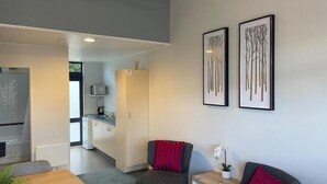 A modern living room with a dining table, two grey armchairs, and a small side table with a plant. There are framed artworks on the wall and a kitchen area in the background. - Academy Motor Inn (Tauranga)