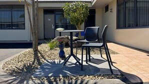 An outdoor seating area with a table and two chairs, surrounded by a small garden with a palm tree and a potted plant. - Academy Motor Inn (Tauranga)