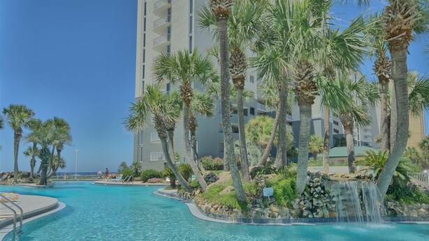 A swimming pool with a waterfall feature, surrounded by palm trees and a multi-story building. - Tower 3-102 Long Beach Resort. Free Beach Chair Service! (Panama City Beach)