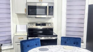 A compact kitchen with white cabinets, a microwave, and an oven. There is a round table with blue upholstered chairs and a marble-patterned tablecloth.