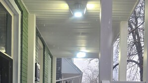 A covered porch with steps leading to a door, white railings, and a view of a building in the background.