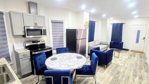 Cocina compacta con electrodomésticos modernos, comedor con sillas azules y pequeño espacio de sala con sofá y cortinas.