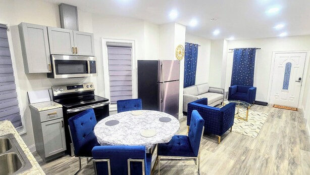A compact kitchen with modern appliances, a dining area with blue chairs, and a small living space with a sofa and curtains.