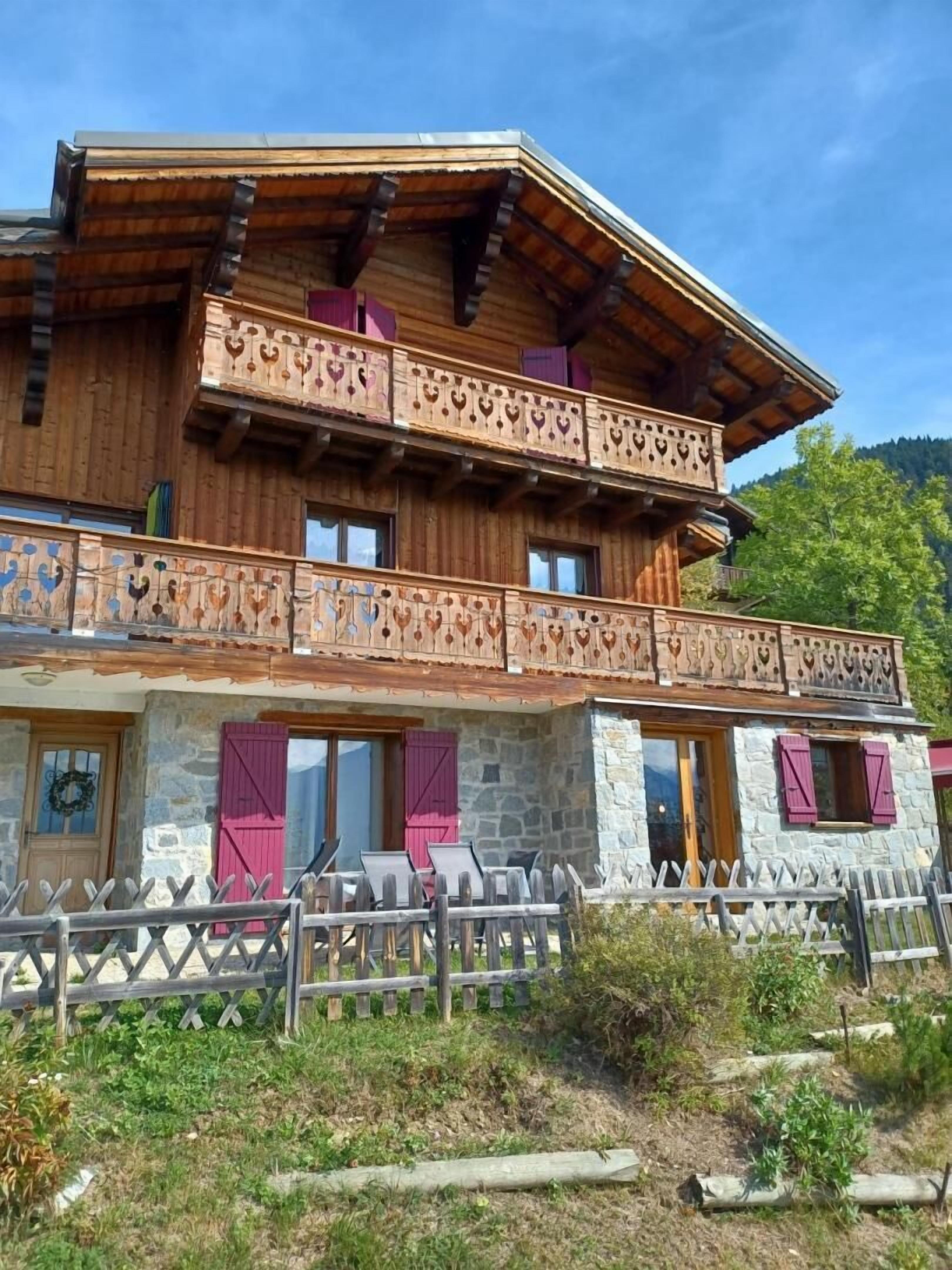 A wooden chalet with a stone facade, balconies with heart-shaped cutouts, and a wooden fence in front.