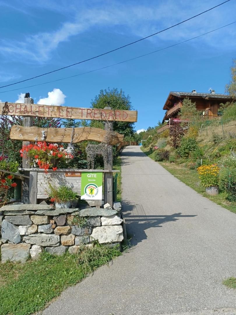 A wooden signpost with 'CHALET TIRETARD' and a 'GITE' sign, a stone wall, and a paved path leading to a wooden building.
