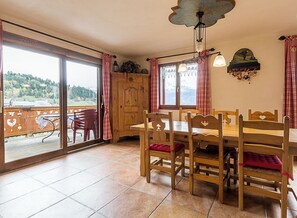 Un'area pranzo con un tavolo e sedie in legno, vista su un paesaggio montuoso attraverso ampie finestre e un balcone con ringhiera.