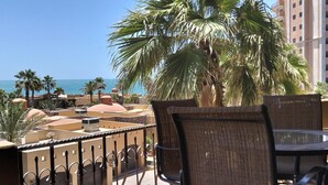 Een balkon met rieten stoelen en een tafel, met uitzicht op een strand met palmbomen en een gebouw op de achtergrond.