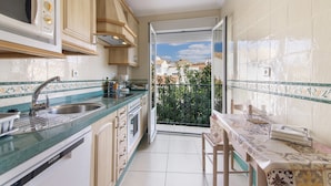 A kitchen with a sink, microwave, and cabinets, leading to a balcony with a view of buildings and greenery.