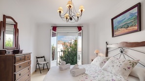 A bedroom with a bed, dresser, chair, chandelier, painting, and a balcony view.