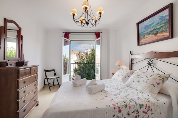 A bedroom with a bed, dresser, chair, chandelier, painting, and a balcony view.