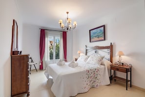 A bedroom with a bed, a wooden nightstand, a desk, a chair, a wardrobe, a chandelier, a window with curtains, and a painting on the wall.
