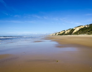Ein Strand mit klarem Himmel, ruhiger See und sandigem Ufer, im Hintergrund Felsen.