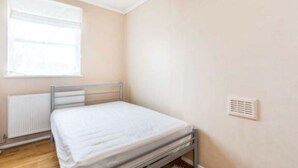 A small, single-bed room with a metal frame, a window with blinds, a radiator, and a wall-mounted air vent.