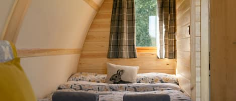 A cozy bedroom with a sloped ceiling, wooden walls, a bed with floral bedding, a pillow with a rabbit design, and a window with plaid curtains.