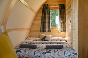 A cozy bedroom with a sloped ceiling, wooden walls, a bed with floral bedding, a pillow with a rabbit design, and a window with plaid curtains. - Rural Ensuite Wooden Cabin With Wood Fired Hot Tub (Tamworth)