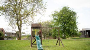 A playground with a slide and swings, surrounded by grass and trees. - Ensuite Wooden Cabin in Rural Staffordshire (Tamworth)