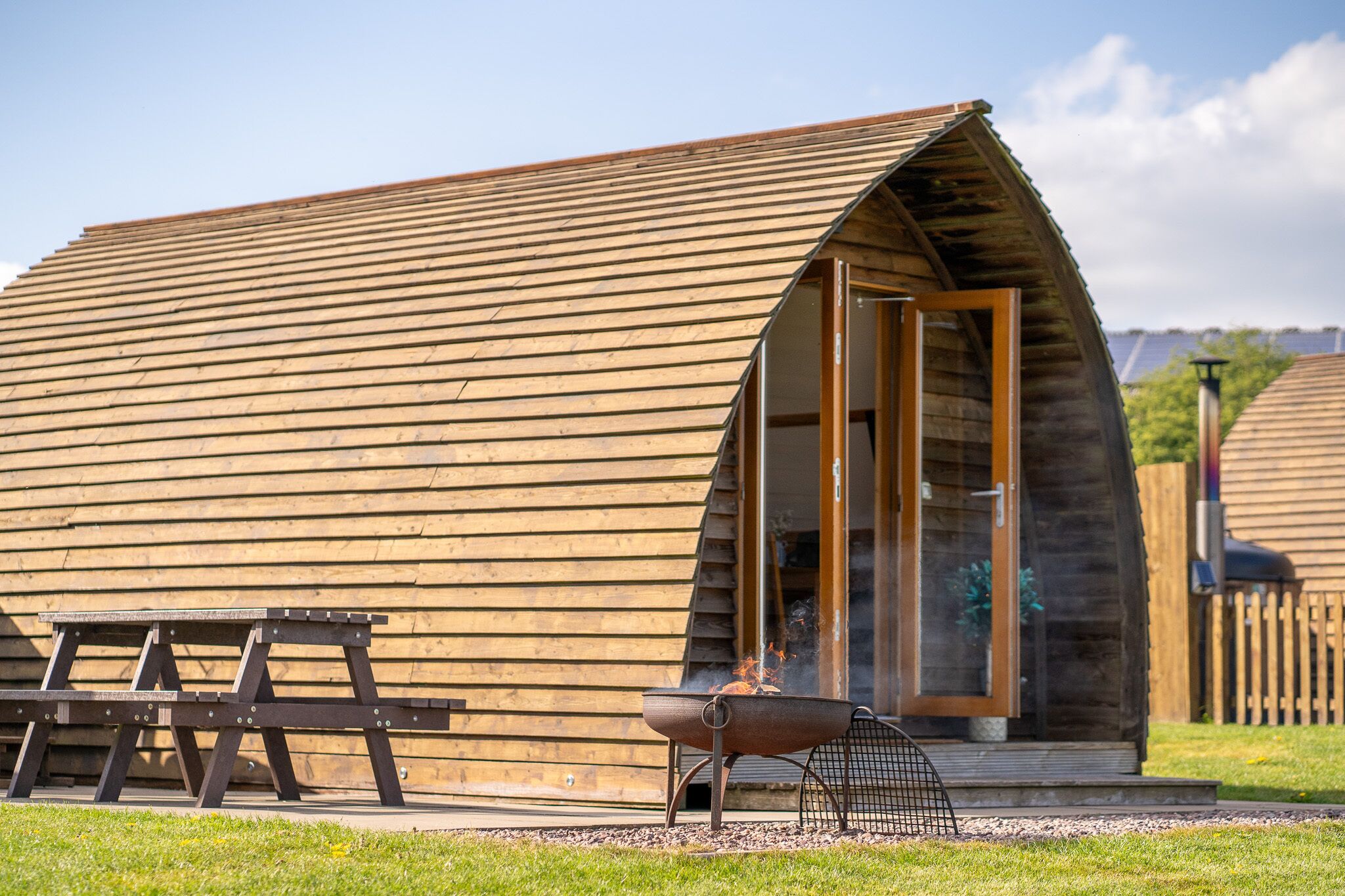 A wooden cabin with a sloped roof, a fire pit, and a picnic table.
