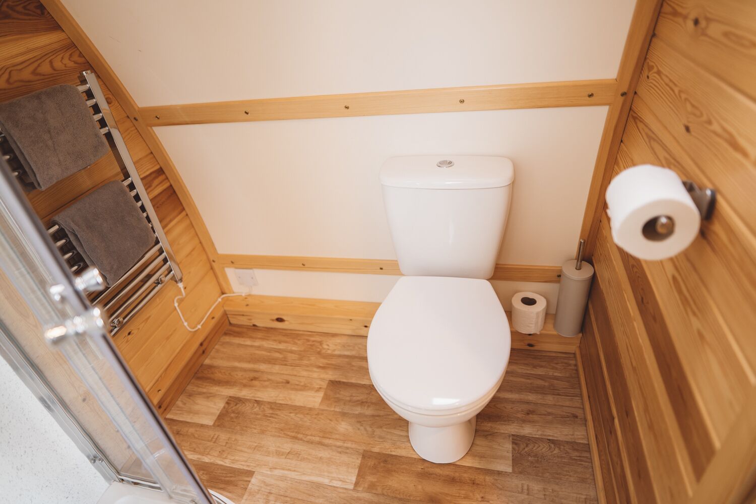 Un baño pequeño con piso de madera, un inodoro blanco, un perchero para toallas y un dispensador de papel higiénico.