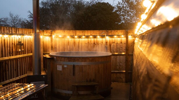 A wooden hot tub with a metal frame, surrounded by a wooden fence and string lights. - Ensuite Wooden Cabin in Rural Staffordshire (Tamworth)